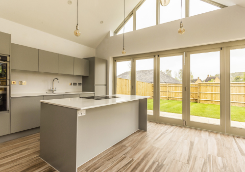 A modern white kitchen with central island and a view looking out over the garden through the large floor-to-ceiling glass patio doors.